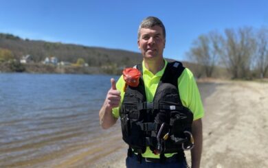 Man wearing a personal flotation device (life jacket) standing along a river bed, smiling and giving a thumbs-up